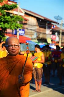 A smiling bold monch, who is wearing a bright orange robe, is taking a Picture of himself and a Parade in the backround with a Selfie-stick.